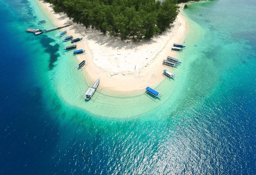 An aerial view of a beach with boats anchored in a circular pattern around a central sandbar, surrounded by clear turquoise waters and a forested island. 1