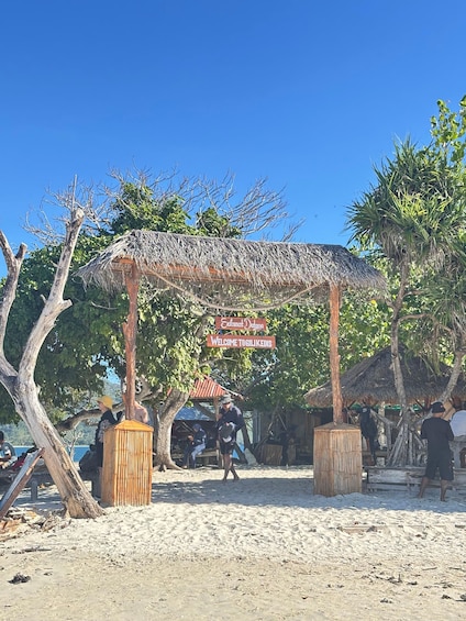 A beach with a thatched-roof structure, a sign reading 'Selamat Darang Welcome to Tigilikedis', and people sitting under trees. 3