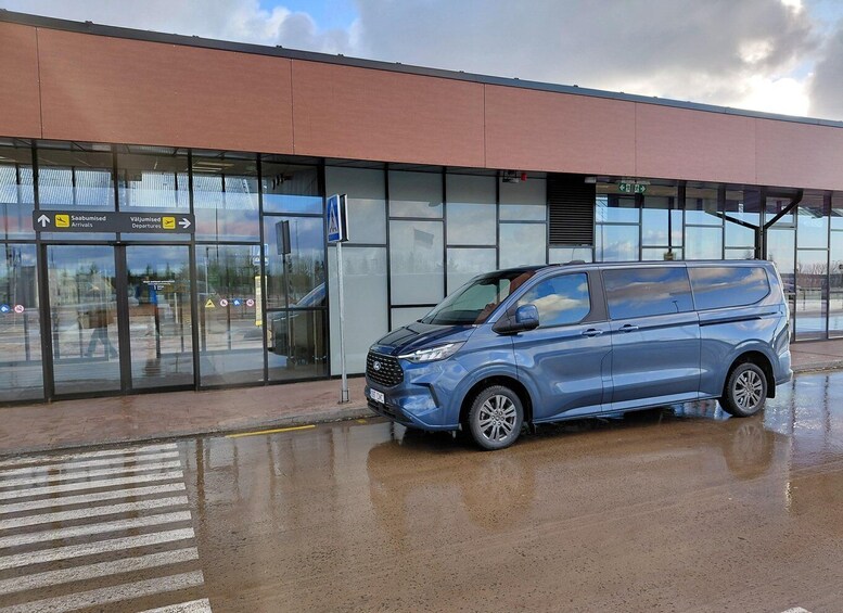 A blue van parked in front of a building with glass doors and a sign indicating arrivals and departures. 4