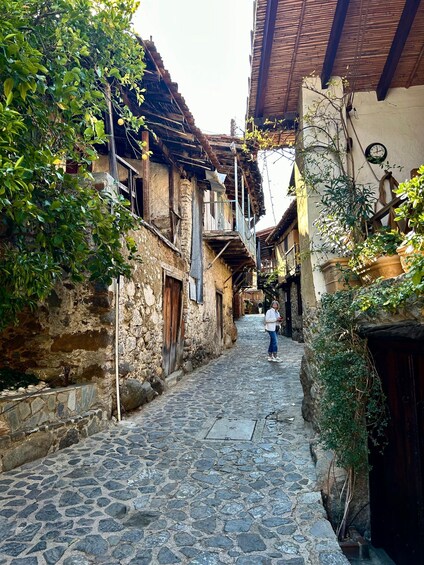 A narrow cobblestone street flanked by traditional stone buildings with wooden balconies and lush greenery. 1