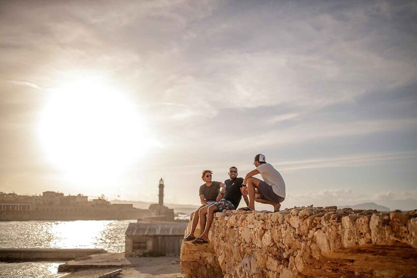 Three people sitting on a stone wall by a waterfront with a lighthouse in the background. 2