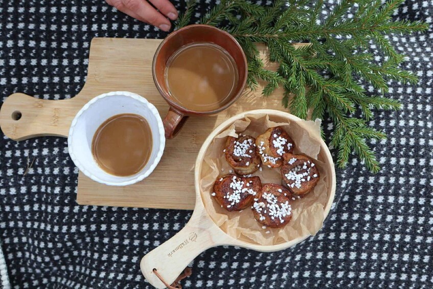A wooden cutting board with a white bowl and a brown mug containing a brown liquid, possibly tea or coffee, and a wooden bowl with cinnamon rolls topped with white sugar. 1