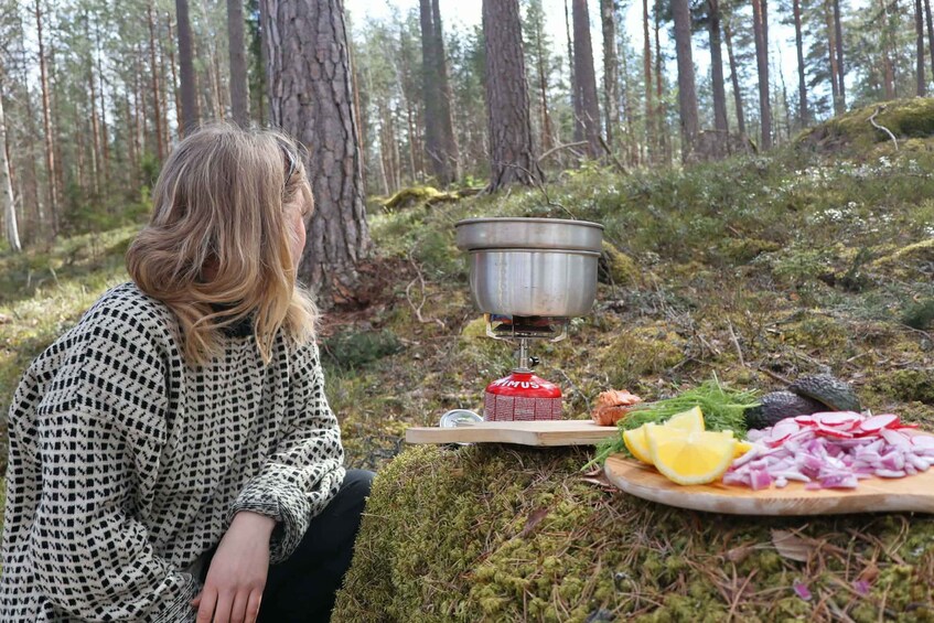 A person sitting by a campfire in a forest with a portable stove, a cutting board with food, and a metal pot. 3