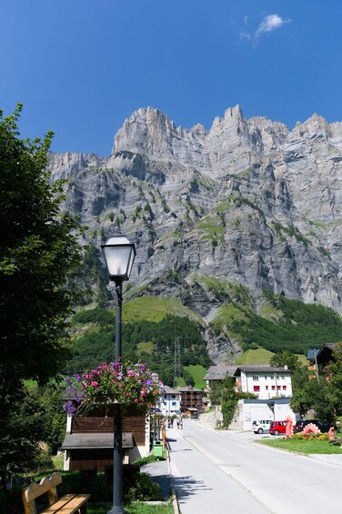 A street lamp with flowers, a bench, a mountain, and buildings in the background. 4
