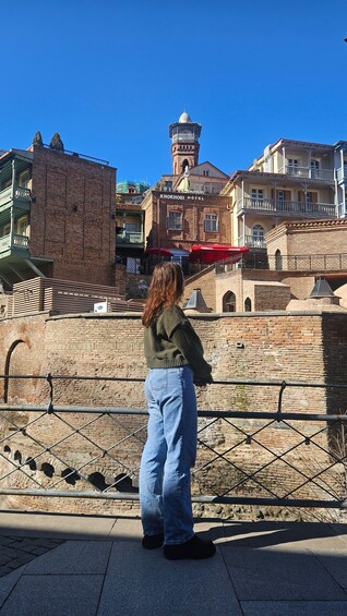 A person standing in front of a historic building with a tower, possibly a hotel, and a brick wall with arches. 3