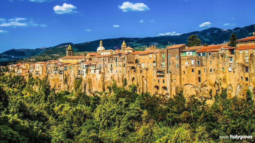A historic town with terracotta-roofed buildings, a prominent church with a bell tower, and lush green hills in the background. 2