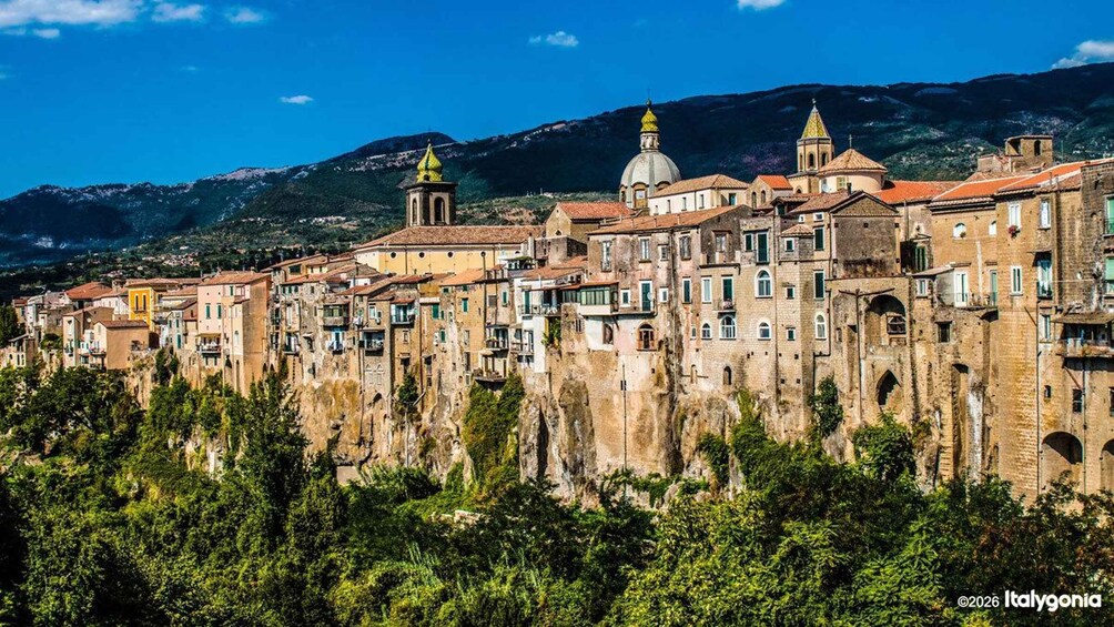 A historic town with terracotta-roofed buildings, a prominent church with a dome, and a mountainous backdrop. 1