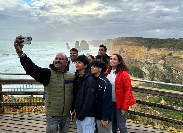 A group of people taking a selfie on a wooden deck overlooking a coastal cliff with distinctive rock formations. 2