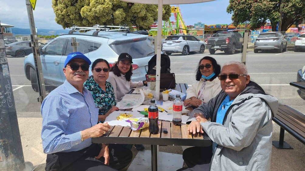 A group of people seated at an outdoor table with various items on it, including a bottle and a bag, in a parking lot with cars and a Ferris wheel in the background. 3
