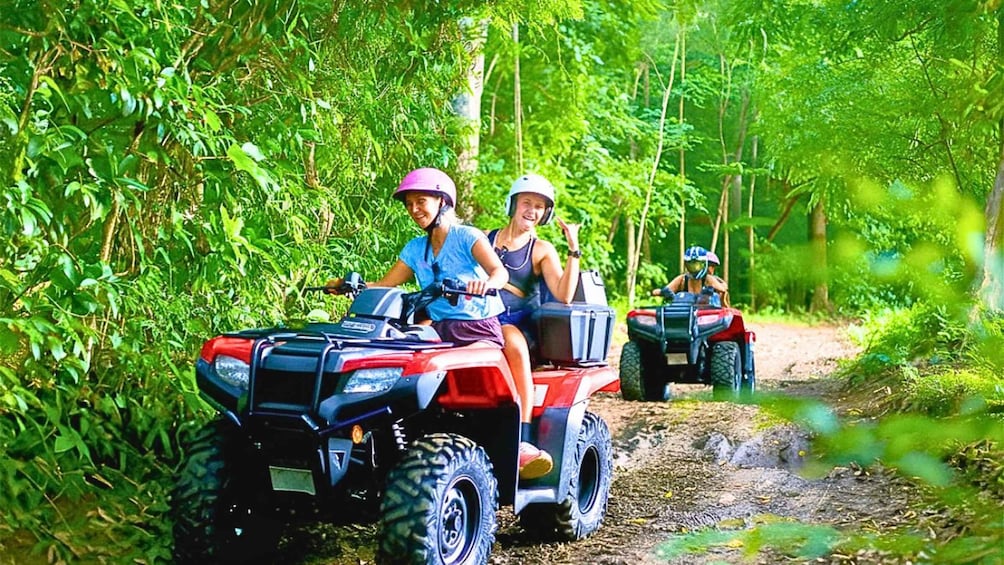 Two people riding ATVs on a dirt path through a forest. 1