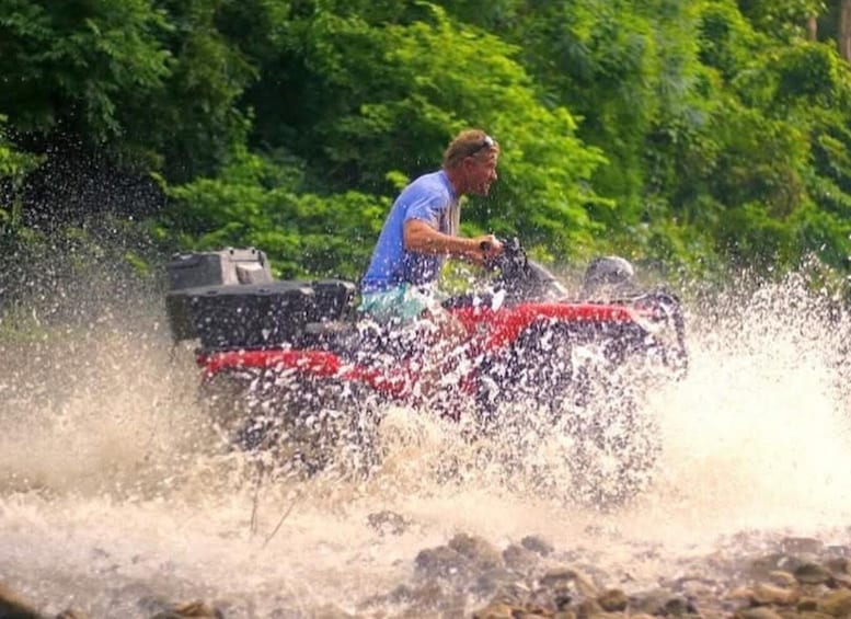 A person riding a red ATV through a body of water, surrounded by greenery. 3