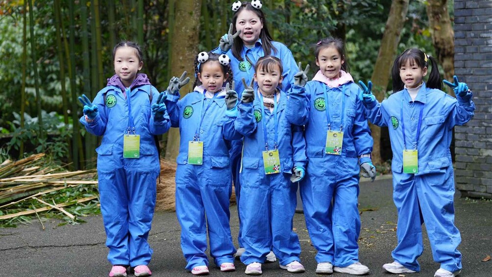 A group of children in blue uniforms with lanyards, standing outdoors with trees and a brick wall in the background. 2