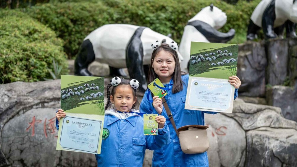 Two individuals holding certificates in front of a panda statue. 1