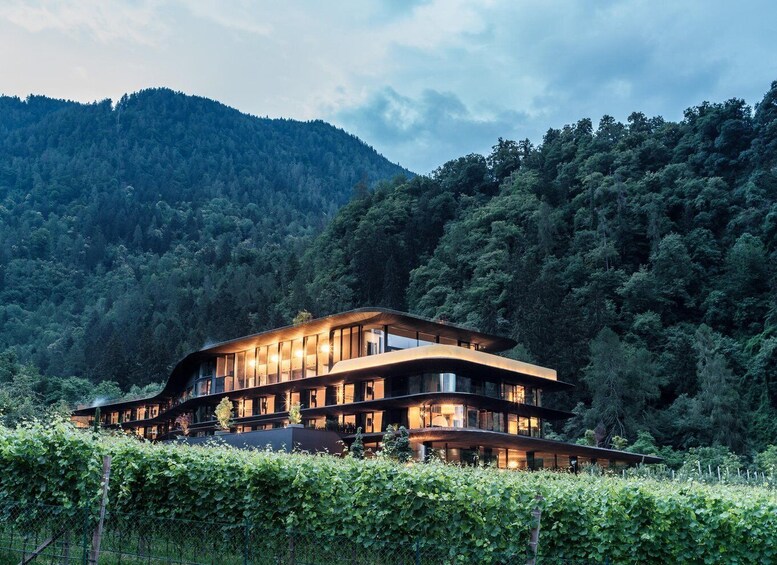 A modern multi-story building with large windows, surrounded by lush greenery and a forested mountain backdrop. 2