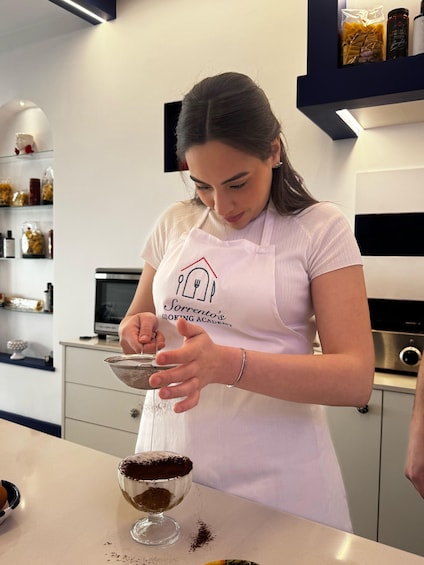 A person in a kitchen, wearing a white apron with a logo, preparing a dessert in a glass bowl. 4