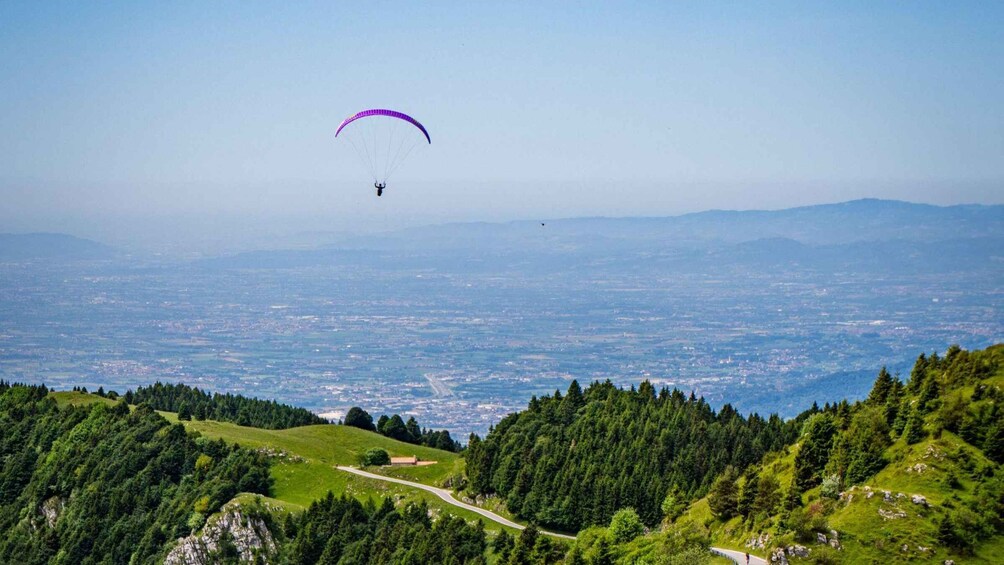 A paraglider with a purple canopy flying over a mountainous landscape with a road and dense forests. 2