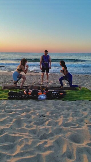 Three people exercising on a beach with a rowing machine and weights in the foreground. 1
