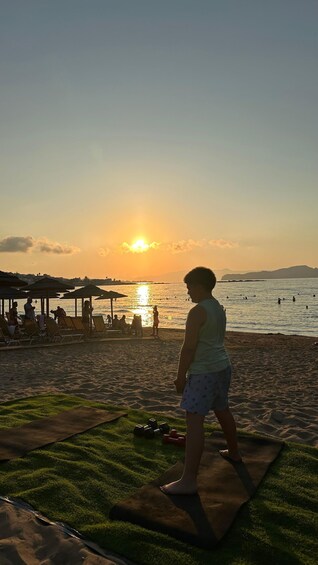 A person on a yoga mat at sunset by the beach with umbrellas and people in the water. 2