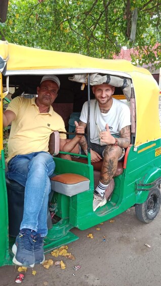 Two people in a yellow and green auto-rickshaw, one wearing a yellow shirt and jeans, the other in a white shirt and shorts, with visible tattoos. 3