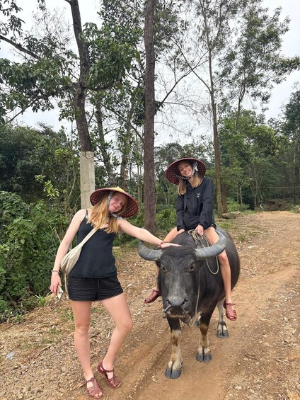 Two people interacting with a large bull on a dirt path surrounded by trees. 2