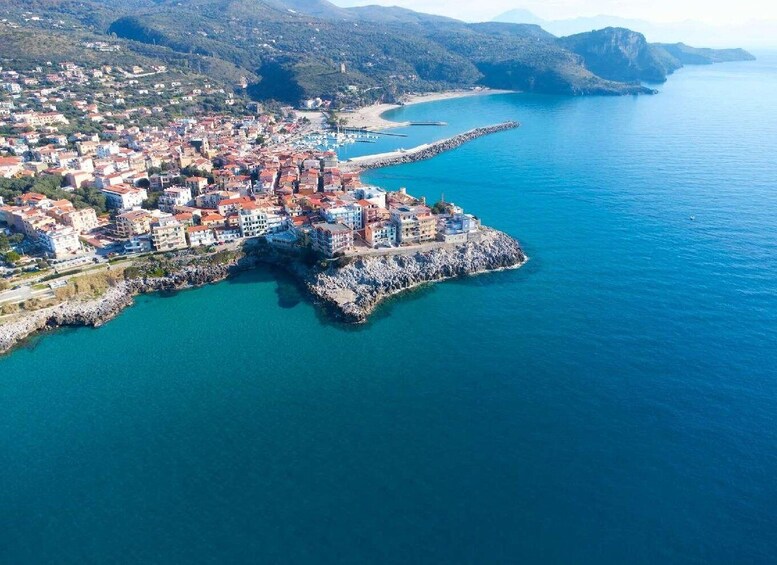 An aerial view of a coastal town with a prominent peninsula, clear blue waters, and surrounding hills. 1