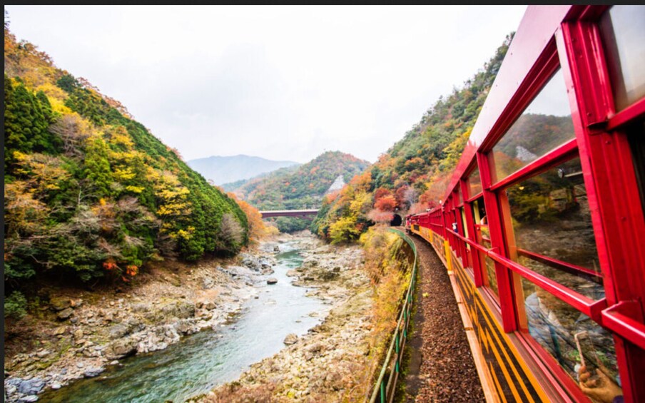 A red train traveling through a valley with a river and autumn-colored trees. 5