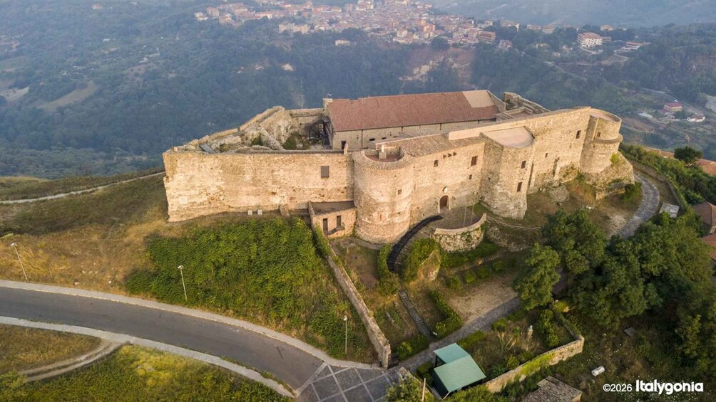 An aerial view of a historic stone castle with a surrounding moat, set against a backdrop of a hilly landscape and a distant town. 1