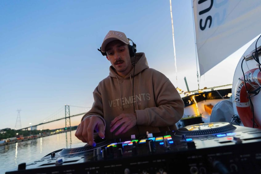 A person operating DJ equipment on a boat with a bridge in the background. 3