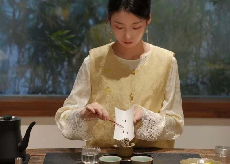 A person in traditional attire preparing tea with a teapot and cups on a table. 3