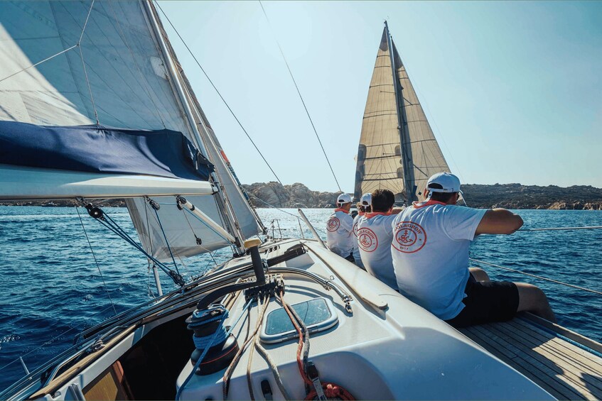 A sailboat with two people wearing matching t-shirts, sailing on a body of water with a rocky coastline in the background. 4