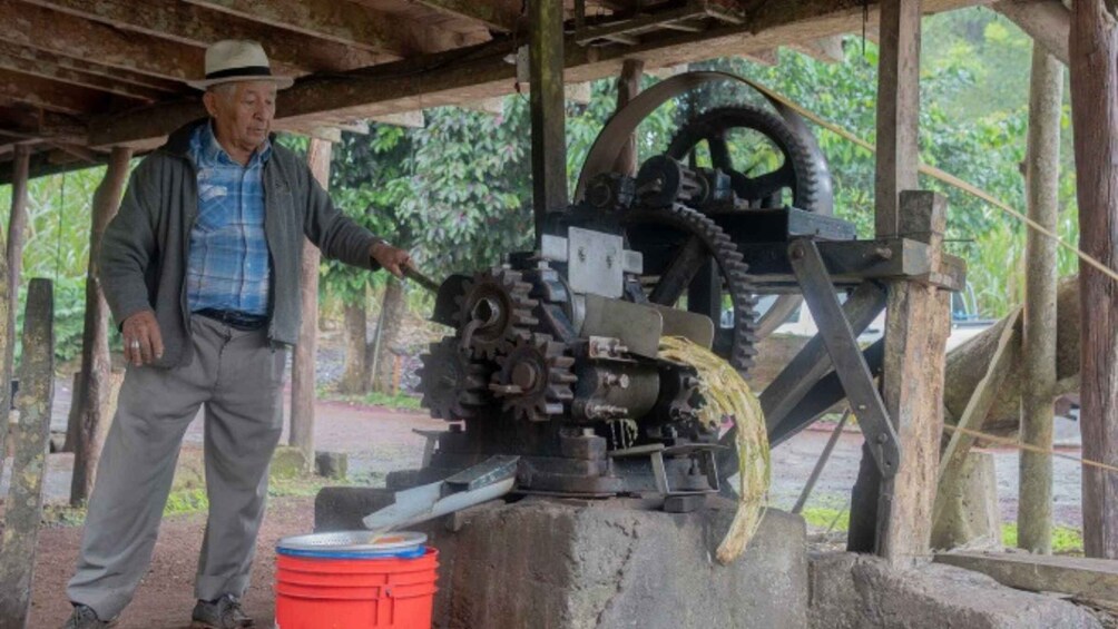 A man standing next to a mechanical device with gears and a wheel, possibly for processing or grinding, under a wooden structure. 3