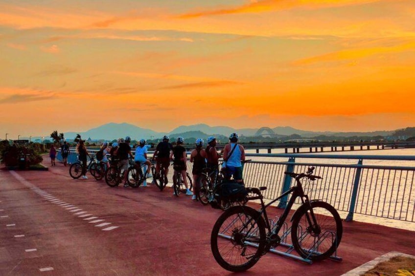 A group of cyclists on a bridge during sunset with a cityscape in the background. 1