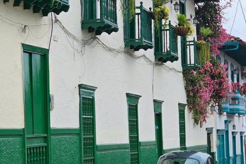 A building with green shutters and white walls, a car parked in front, and hanging plants. 1