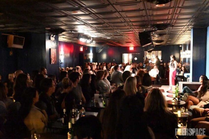 A crowded bar with people seated and standing, a stage with a performer, and a dark interior with visible ceiling beams. 4