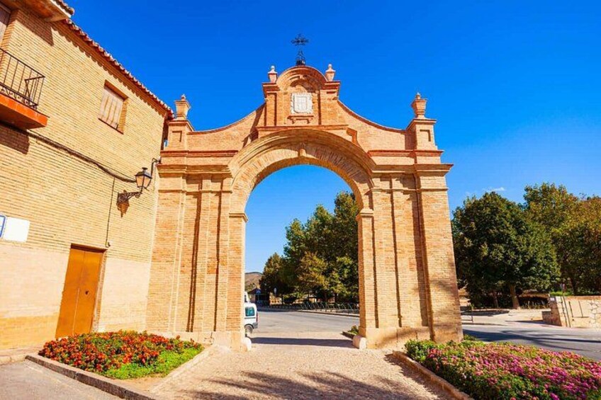 A stone archway with a clock, surrounded by flowers and buildings. 5