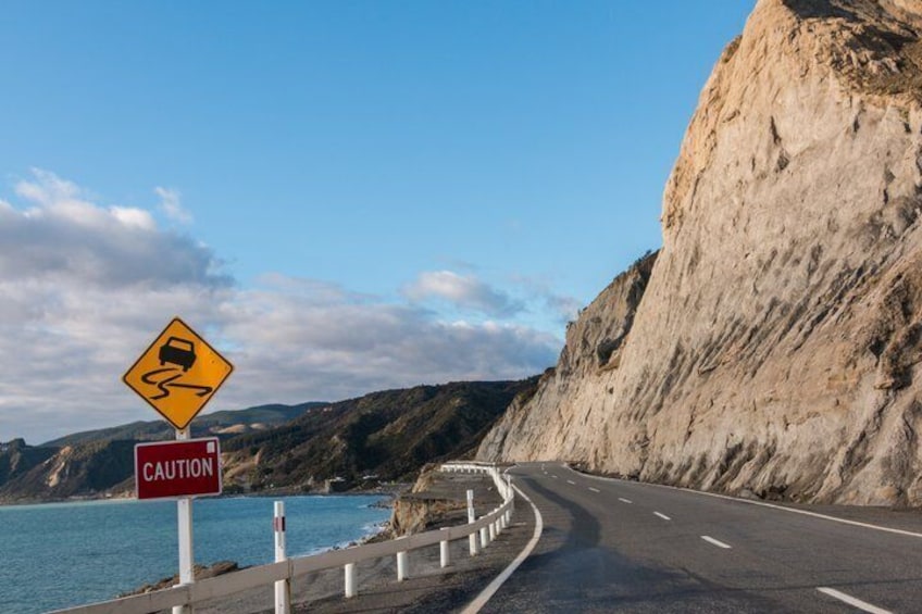 A coastal road with a caution sign for slippery conditions, a red 'CAUTION' sign, and a clear blue sky. 2