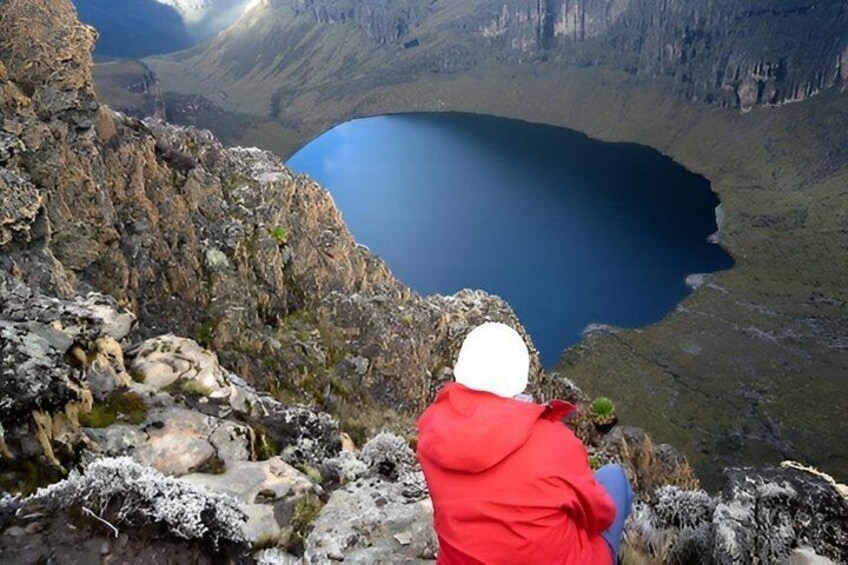 A person in a red jacket sitting on a rocky outcrop overlooking a deep blue lake surrounded by rugged terrain. 1