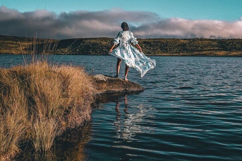 A person standing on a rock by a calm body of water with grass and hills in the background. 4