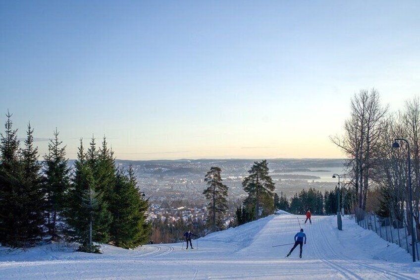 A snowy ski slope with skiers, a clear sky, and a distant cityscape. 2
