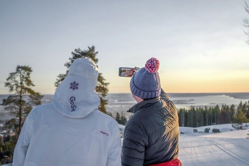 Two people in winter clothing, one wearing a white jacket with a logo, and the other in a gray jacket with a red pom-pom hat, overlooking a snowy landscape with trees and a distant view of a body of water. 3
