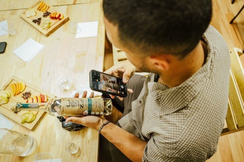 A person holding a smartphone and a bottle of liquor at a wooden table with various food items and glasses. 5