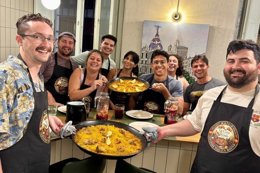 A group of people around a table with a large pan of food, likely a dish with rice and meat, and a pitcher of a red sauce. 5