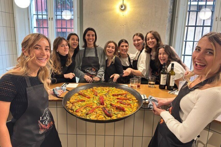 A group of people around a large pan of food, possibly a paella, in a kitchen setting. 1