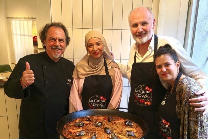 Four people in a kitchen, three wearing aprons with 'La Cuina Nicolas' text, and a large pan of food in the foreground. 2