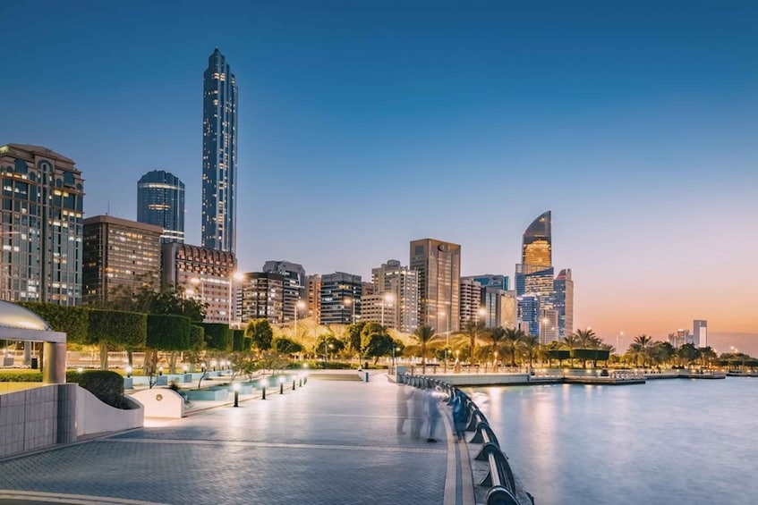 A city skyline at dusk with illuminated buildings, a waterfront promenade, and palm trees. 2