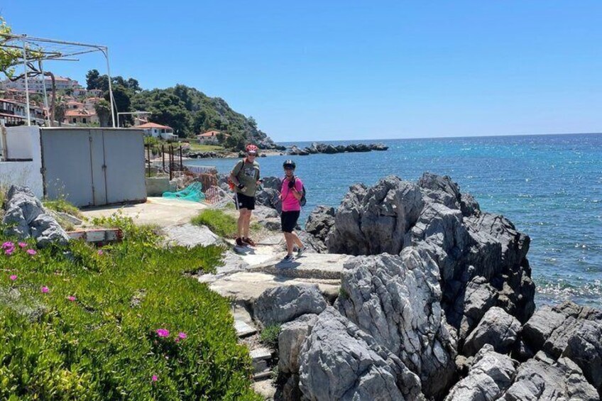 Two people walking on a rocky path by the sea, with buildings and greenery in the background. 4
