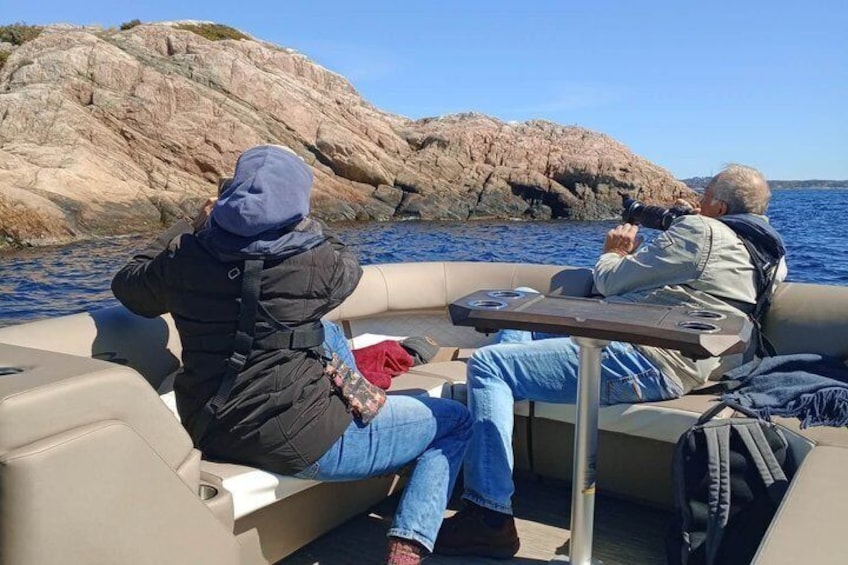 Two people on a boat, one taking a photo, with rocky coastline in the background. 3