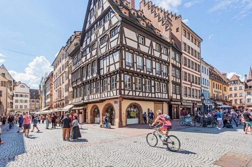 A cobblestone square with historic buildings, a cyclist, and people walking. 4