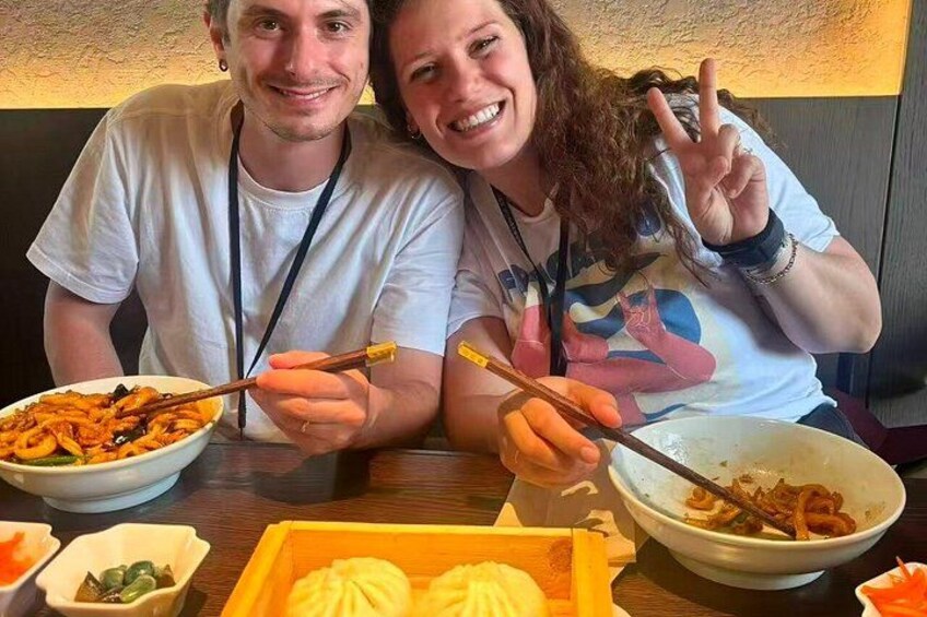 Two people sitting at a table with bowls of food, chopsticks, and a box of dumplings. 2
