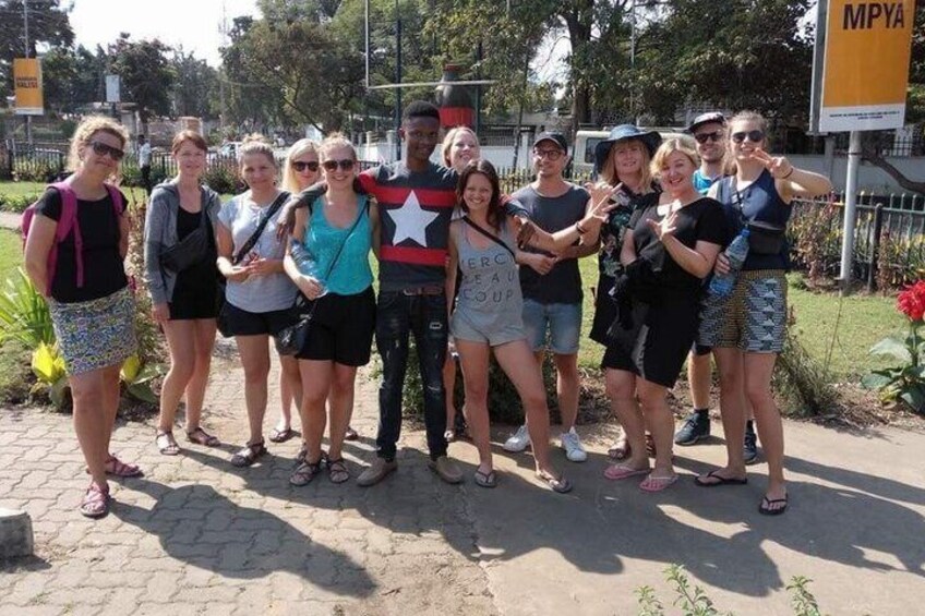 A group of people standing on a paved path with greenery and a sign in the background. 1